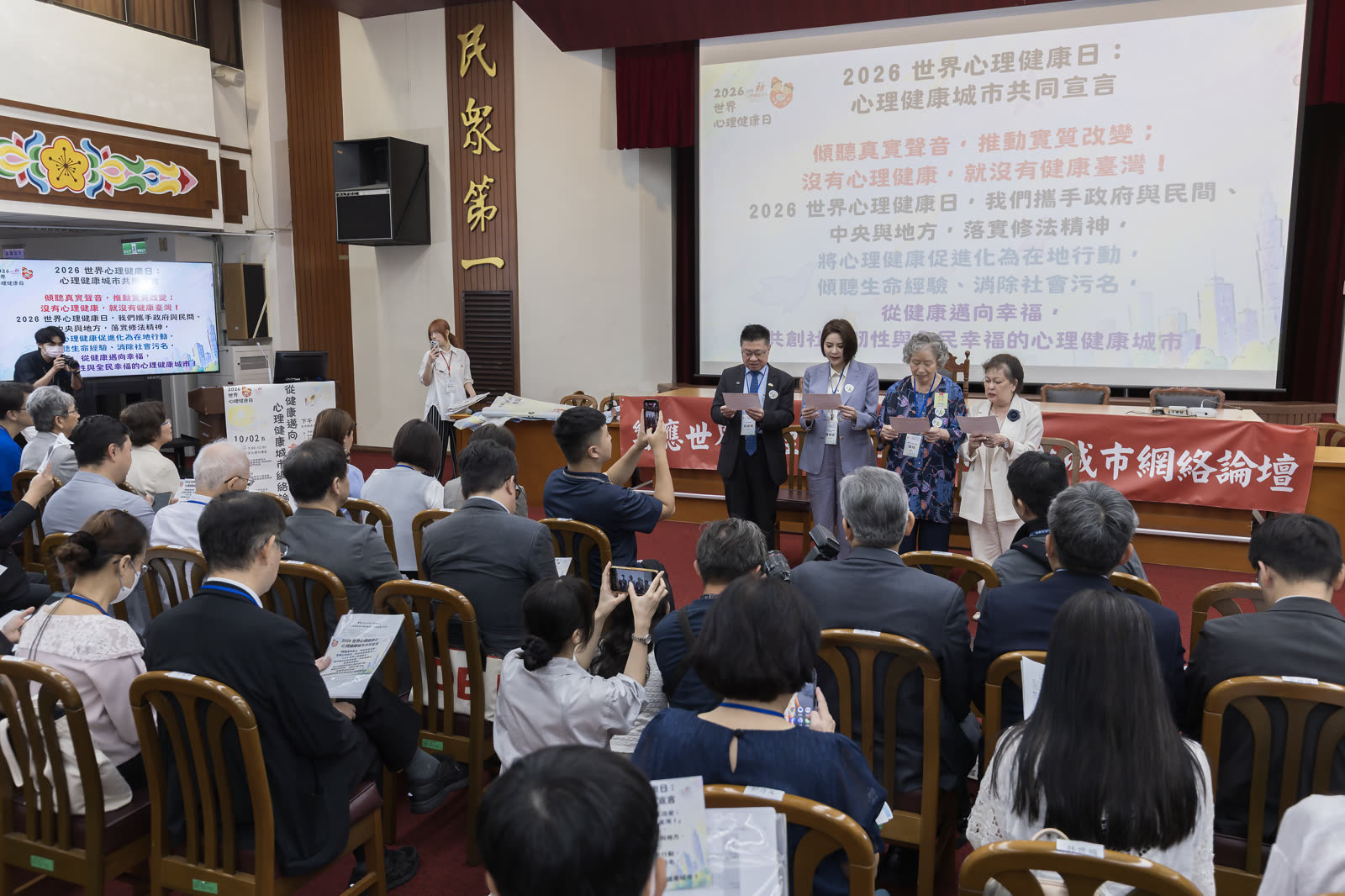 宣言發表現場，台上數人手持紙張面向台下，投影幕顯示心理健康城市共同宣言